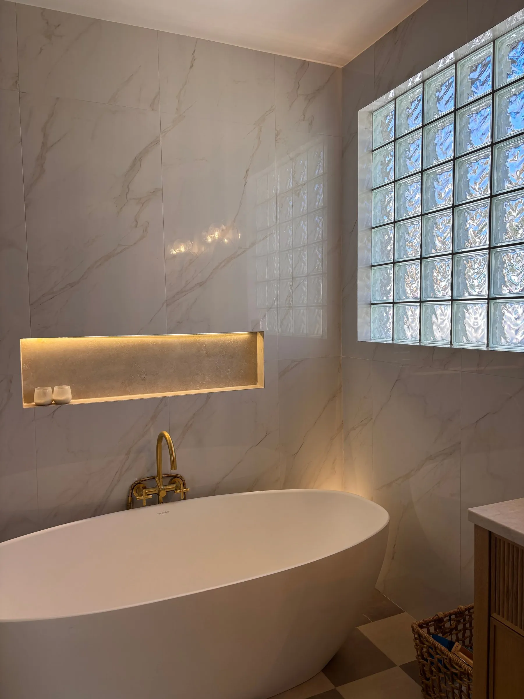 Freestanding oval tub below a glass block window, with a brass floor-standing filler and a lit marble niche