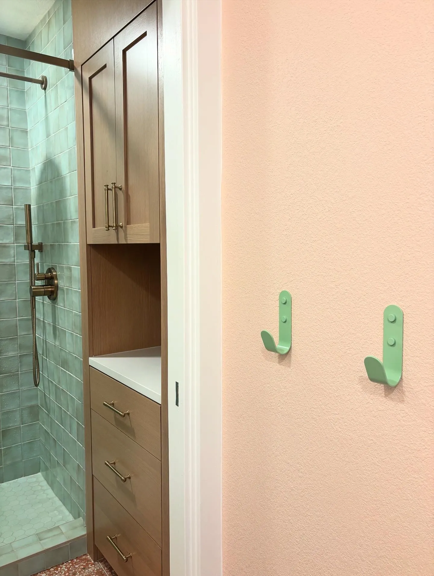 Mint green wall hooks on a blush pink wall beside a tall white oak linen cabinet, the sea-green zellige shower through the doorway