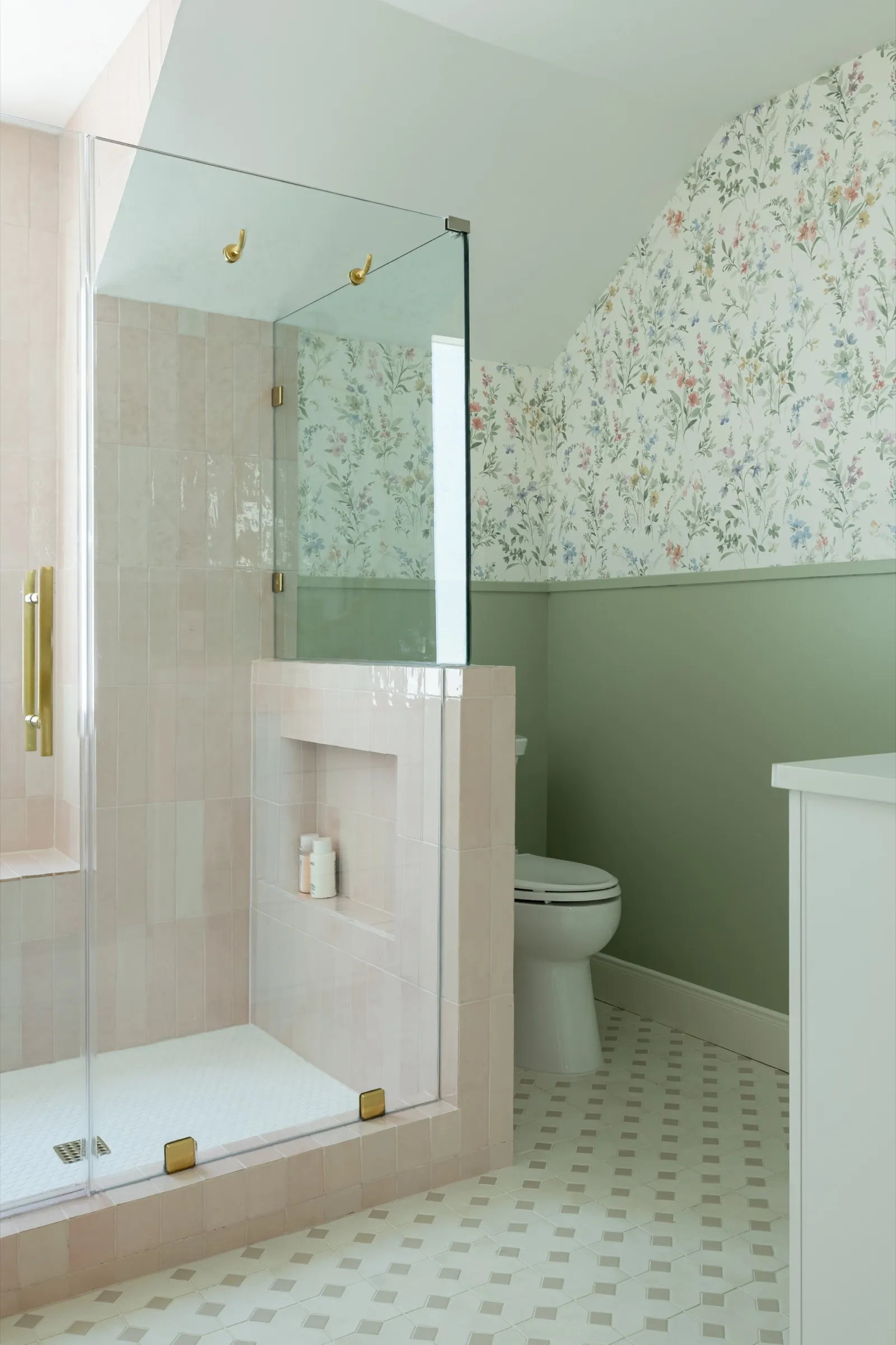 Bathroom with a glass shower lined in pink zellige tile, sage green walls below wildflower paper and a patterned tile floor