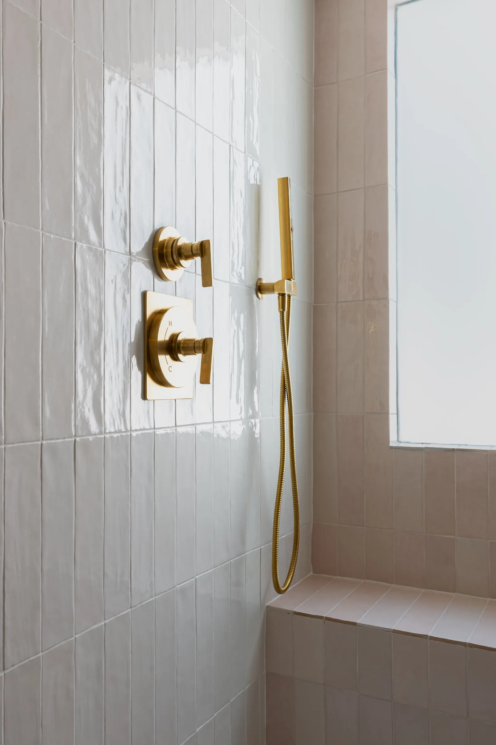 Brass shower valves and handset on white tile above a pink tiled bench