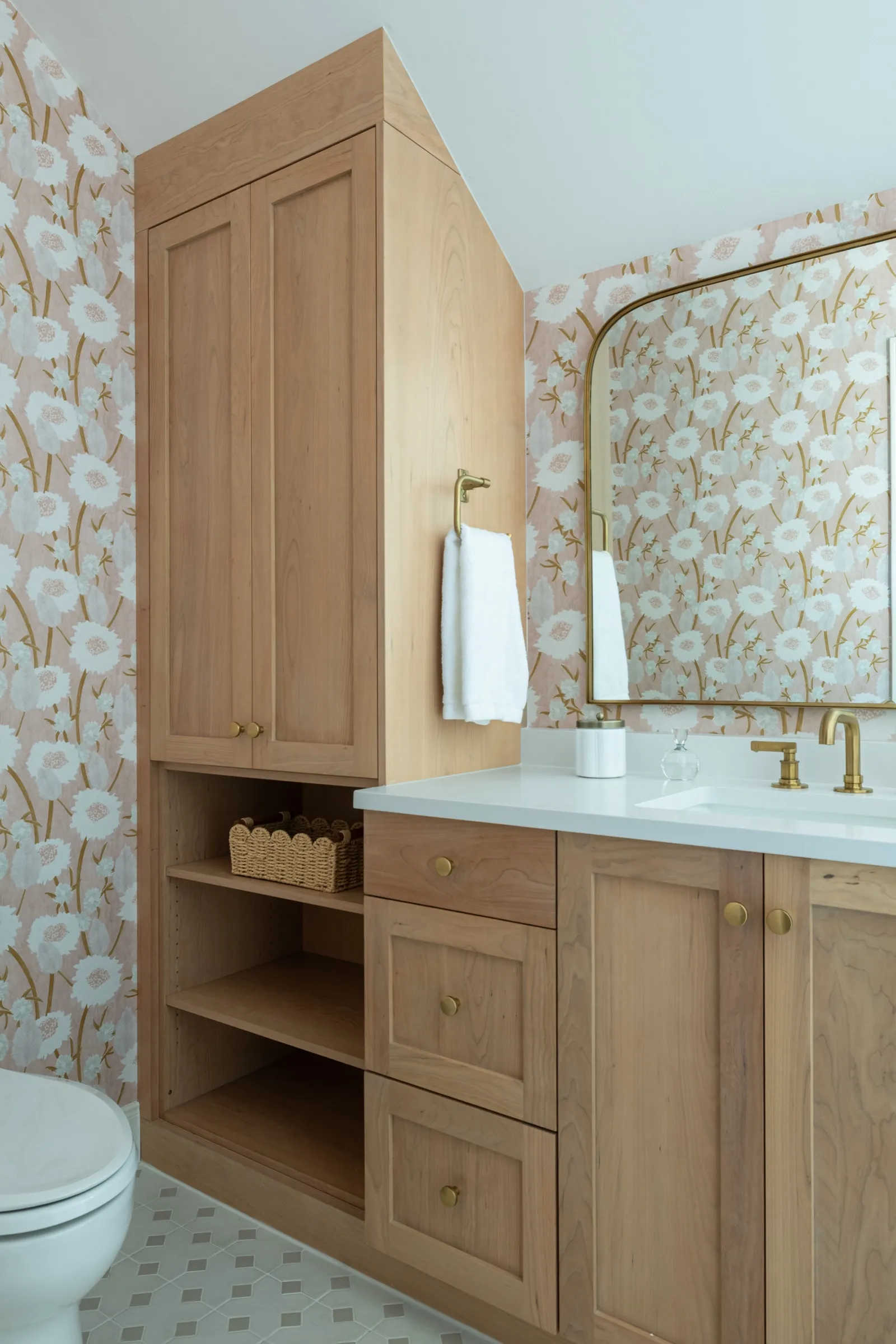 A white oak vanity and tall linen cabinet against blush poppy wallpaper, an arched brass mirror above