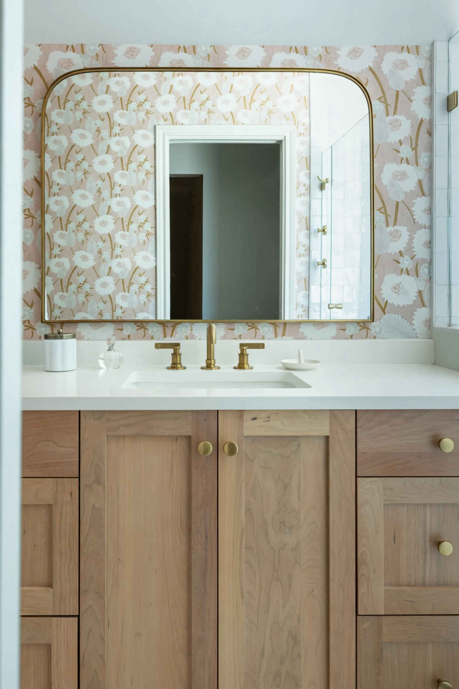 White oak vanity with a brass tap and an arched mirror reflecting the doorway, blush poppy paper behind