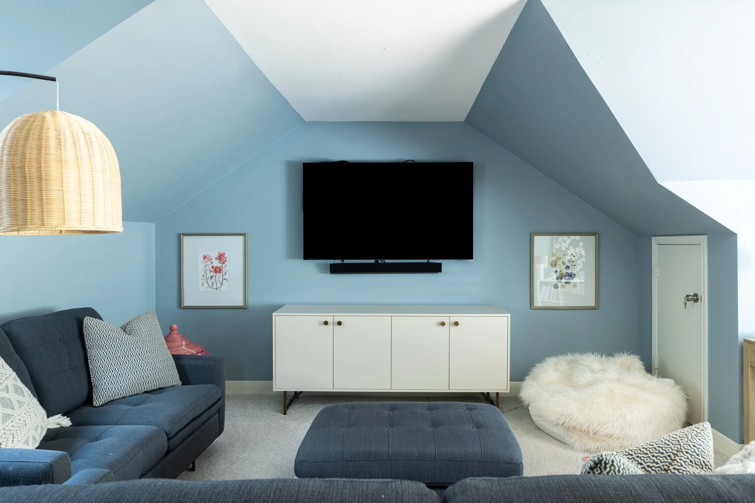 The family room square on: television above a white sideboard, framed prints either side and a navy ottoman